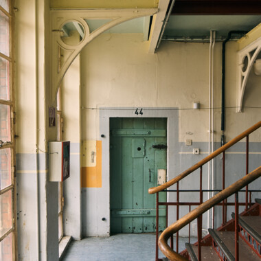 Das Bild zeigt Innere eines älteren Gefängnisgebäudes: ein Flur mit einer massiven, grün gestrichenen Zellentür mit der Nummer 44. Links fällt Tageslicht durch große Fenster herein, rechts führt eine Metalltreppe nach oben. Die Wände sind in hellen, leicht abgenutzten Farben gestrichen, und Rohre verlaufen sichtbar entlang der Wände.